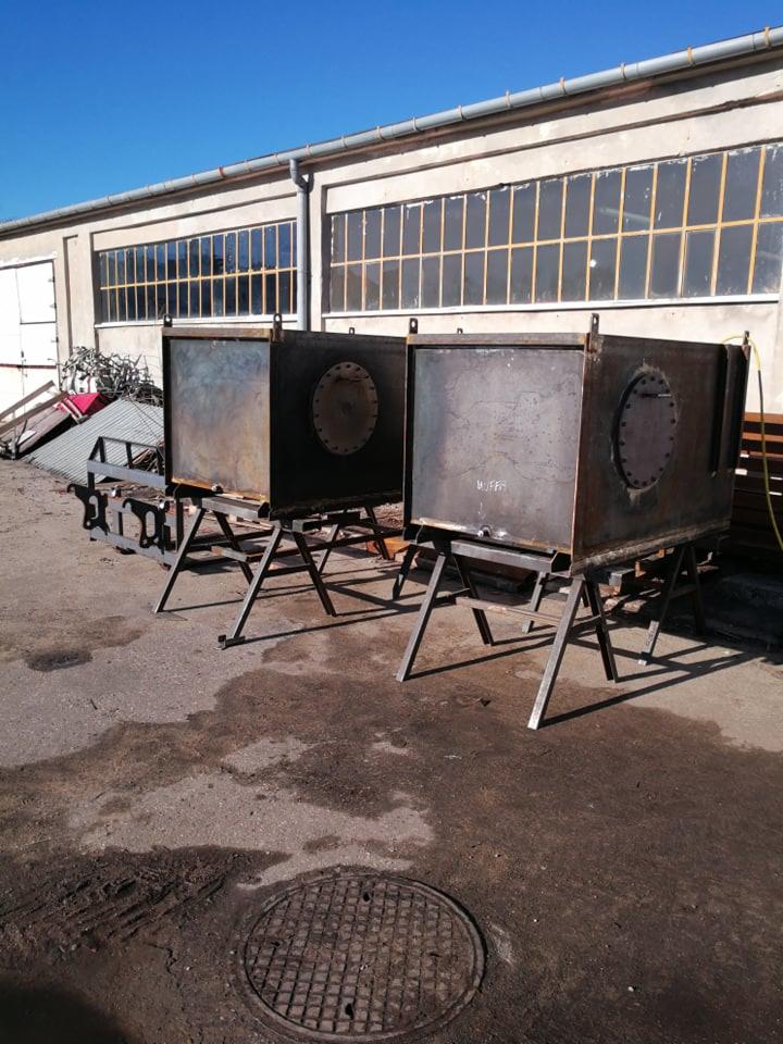 Dwa surowe, metalowe zbiorniki na spawanych konstrukcjach wsporczych, stojące na zewnątrz hali z rzędem okien, widoczne ślady spawania i obróbki metalu.