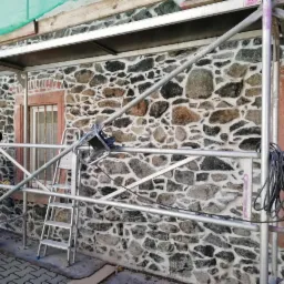 Renowacja kamiennej elewacji budynku z użyciem rusztowania, widoczny pracownik w trakcie czyszczenia, ściana z nieregularnych kamieni w odcieniach szarości i brązu.