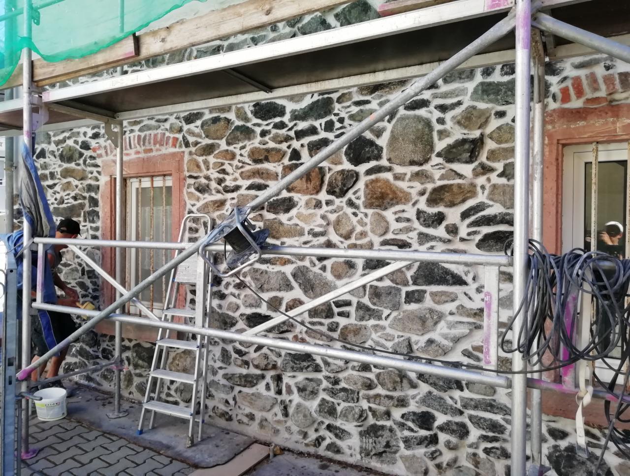 Renowacja kamiennej elewacji budynku z użyciem rusztowania, widoczny pracownik w trakcie czyszczenia, ściana z nieregularnych kamieni w odcieniach szarości i brązu.