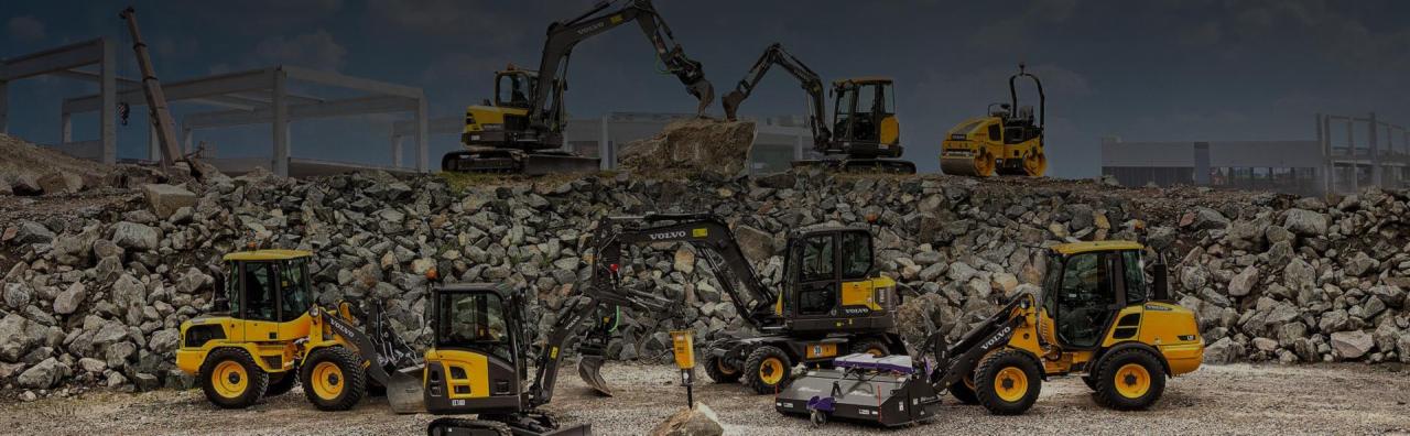 Żółte koparki i walec Volvo na placu budowy z gruzem i konstrukcją budynku w tle.