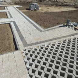 Fragment ścieżki z jasnej kostki brukowej z ciemnymi obrzeżami, przylegający do powierzchni z ażurowych płyt betonowych, widoczne przygotowane podłoże i stosy kostki w tle.