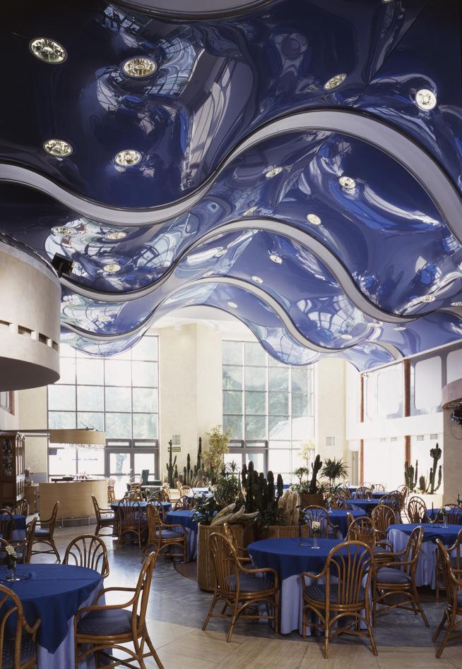 Błyszczący, ciemnoniebieski sufit napinany z wbudowanymi punktowymi lampami, odbijający wnętrze restauracji z drewnianymi krzesłami i stołami nakrytymi niebieskimi obrusami, oraz dużymi oknami w tle.