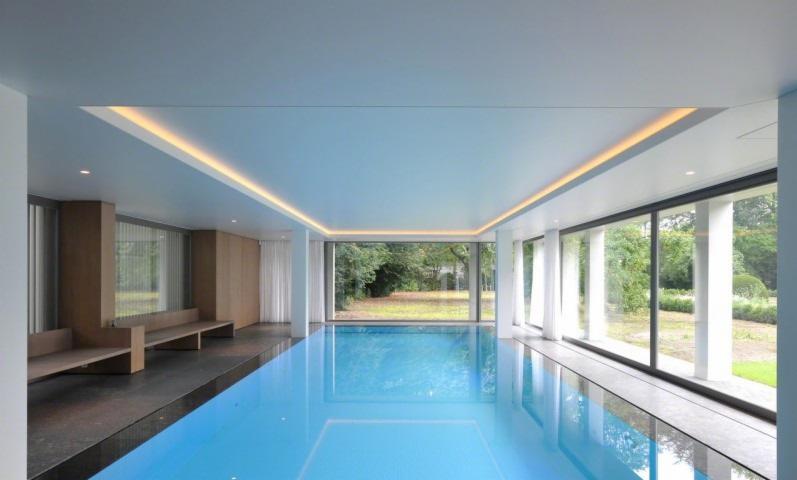 Kryty basen z podświetlanym, gładkim sufitem w kolorze jasnego błękitu, otoczony panoramicznymi oknami z widokiem na zieleń.