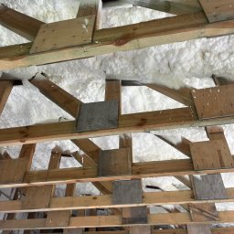 Anibud - Ocieplenie poddasza pianką na drewnianej konstrukcji dachu. Widoczne deski i biała pianka izolacyjna wypełniająca przestrzenie.