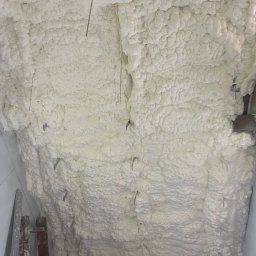 Anibud - Ocieplony poddasze pianką PUR. Widoczne metalowe elementy mocujące i drabina oparta o ścianę. Tekstura pianki przypomina gęstą, białą chmurę.