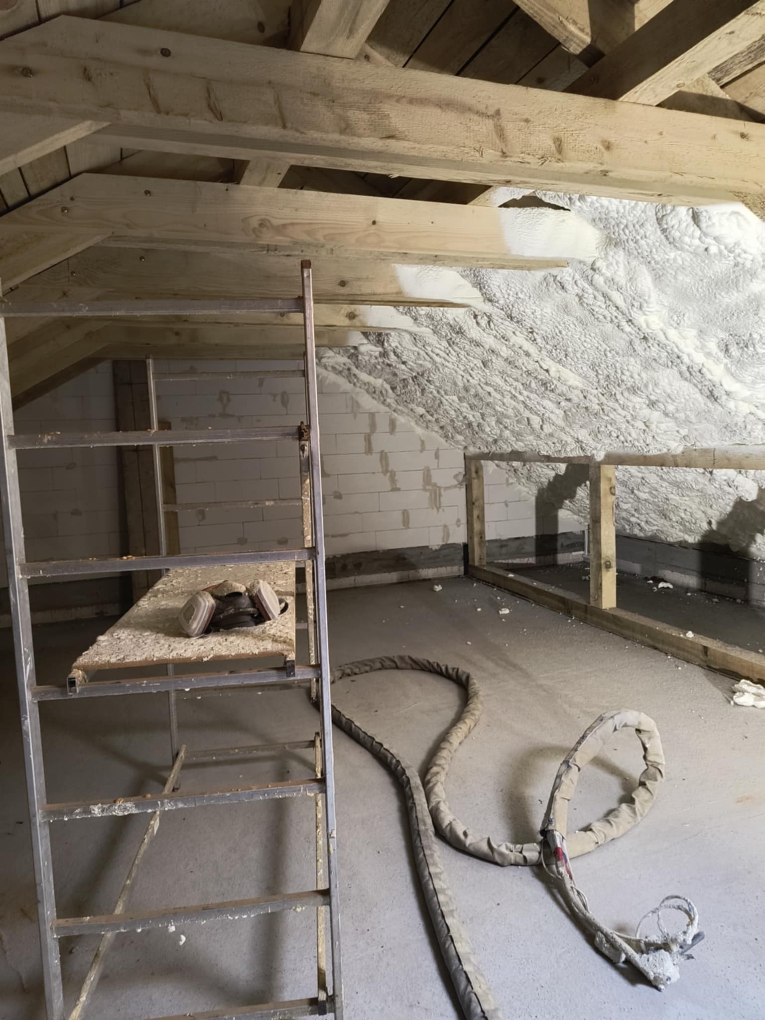 Poddasze po natrysku pianki PUR, widoczne drewniane elementy konstrukcyjne, drabina i węże do aplikacji. Ocieplenie wykonane na bloki betonowe.