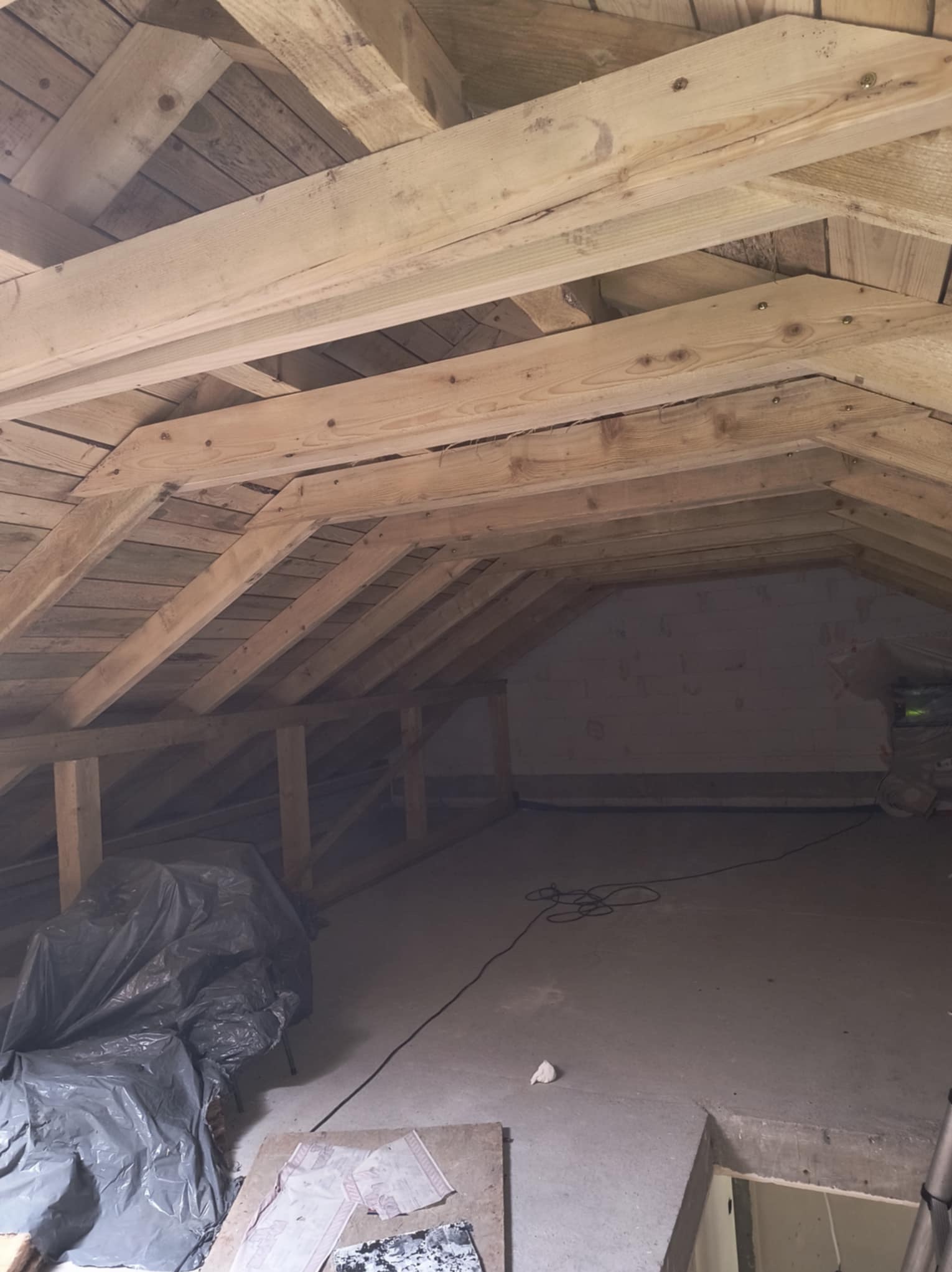 Poddasze z drewnianą konstrukcją więźby dachowej przed ociepleniem. Widoczne belki, deski i przygotowane materiały budowlane. Surowy stan poddasza.
