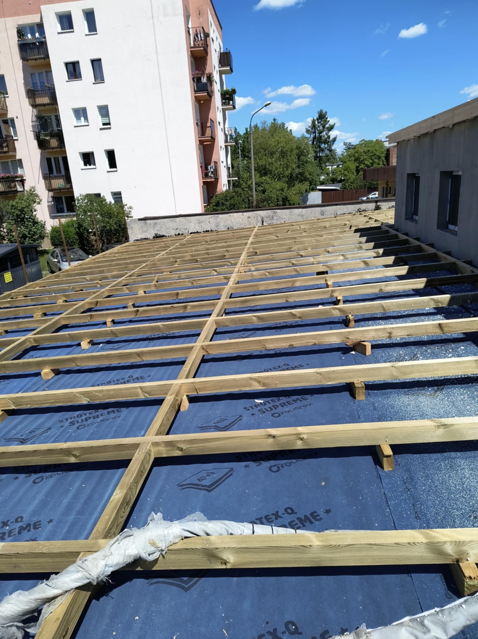 Konstrukcja dachu pod ocieplenie, widok z góry, na tle zabudowań w Żyrardowie. Drewniane belki na membranie. Etap przygotowania do izolacji.