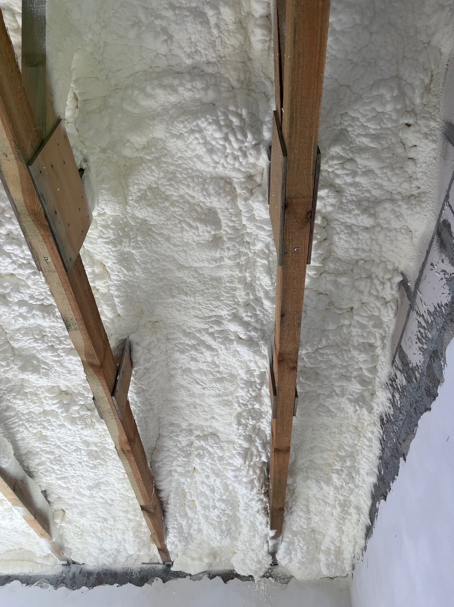 Izolacja poddasza pianką PUR, widoczne drewniane krokwie i biała, spieniona warstwa izolacyjna, szczelnie wypełniająca przestrzeń między elementami konstrukcyjnymi.