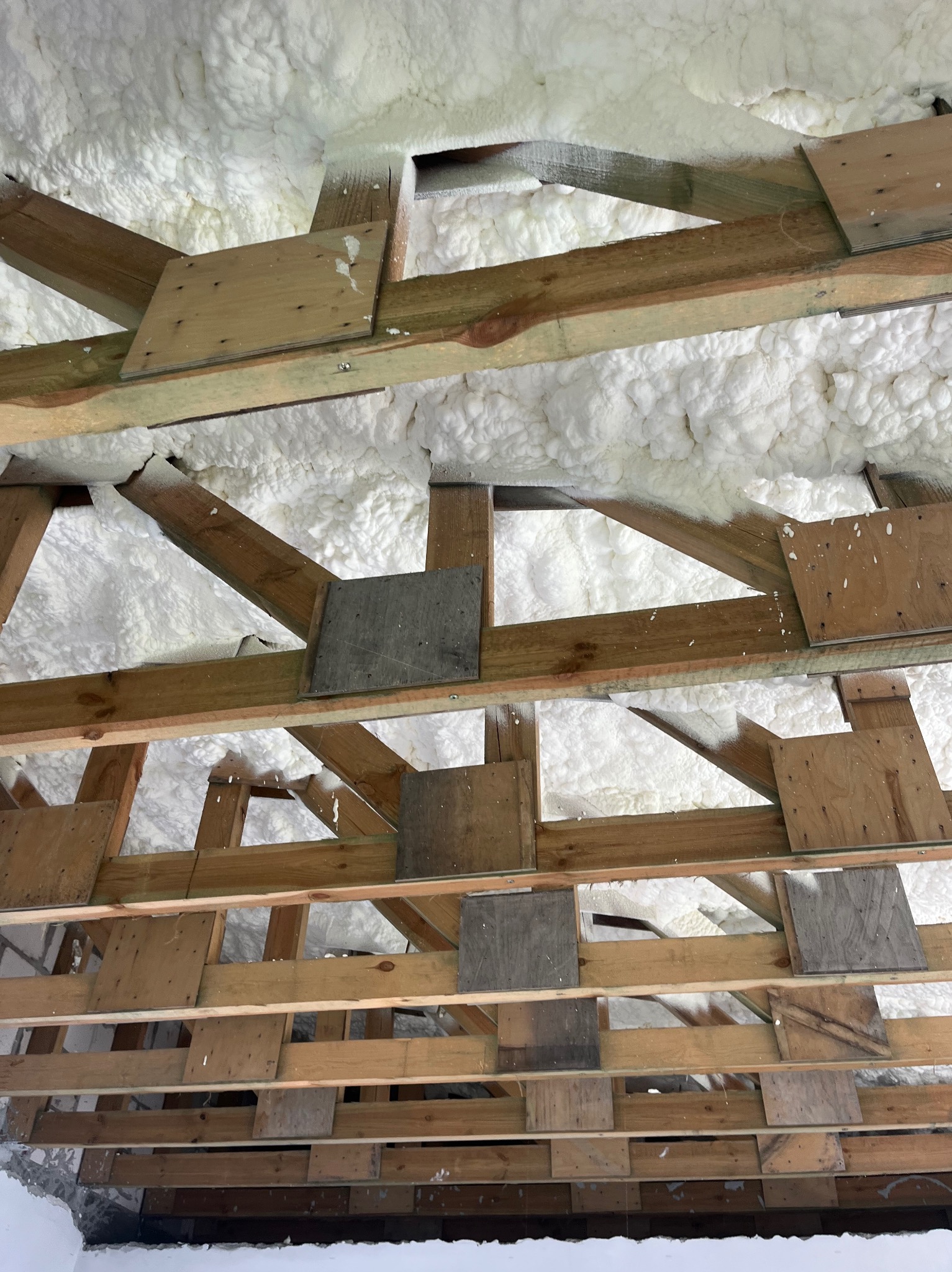 Ocieplenie poddasza pianką na drewnianej konstrukcji dachu. Widoczne deski i biała pianka izolacyjna wypełniająca przestrzenie.