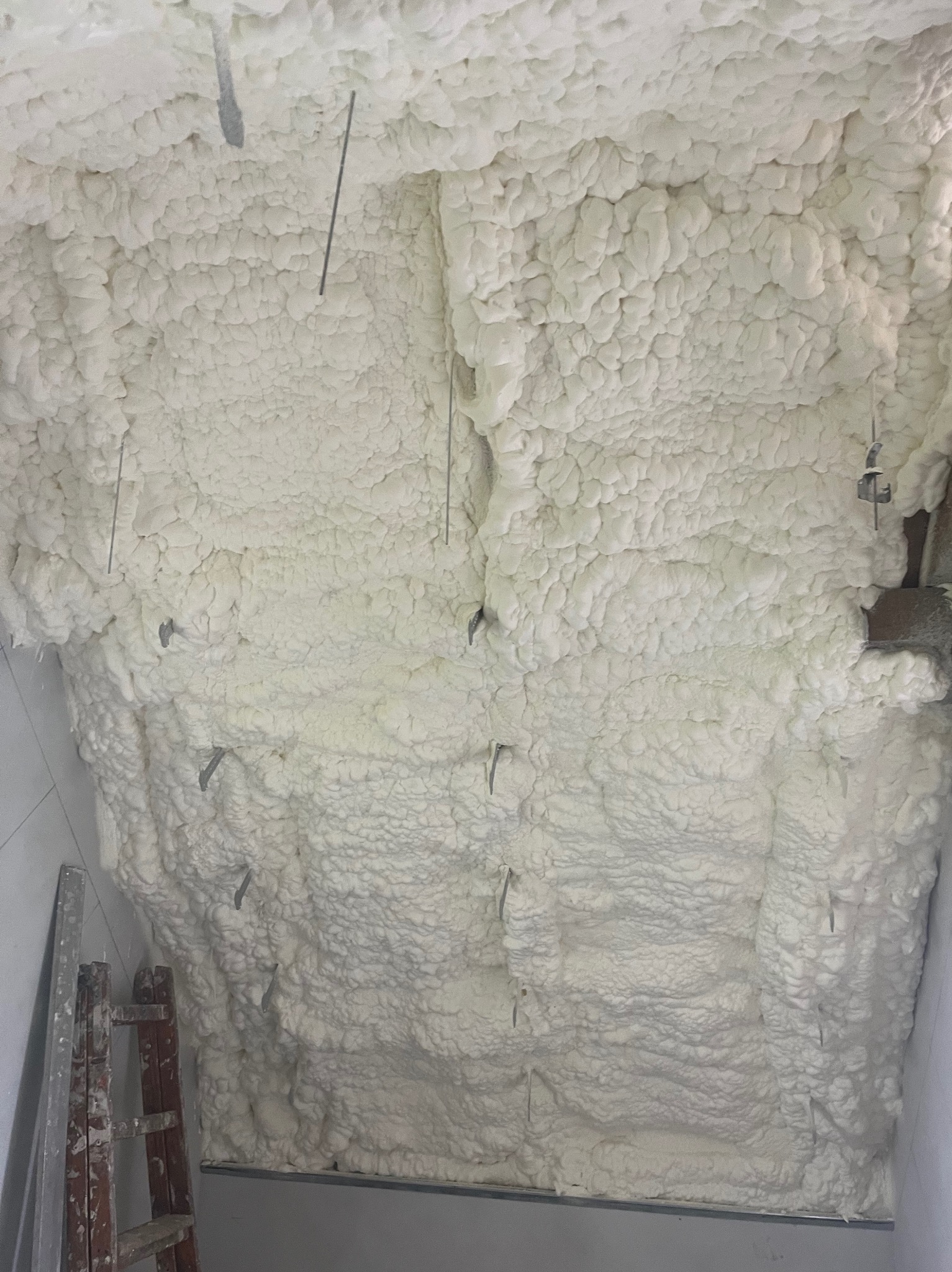 Ocieplony poddasze pianką PUR. Widoczne metalowe elementy mocujące i drabina oparta o ścianę. Tekstura pianki przypomina gęstą, białą chmurę.