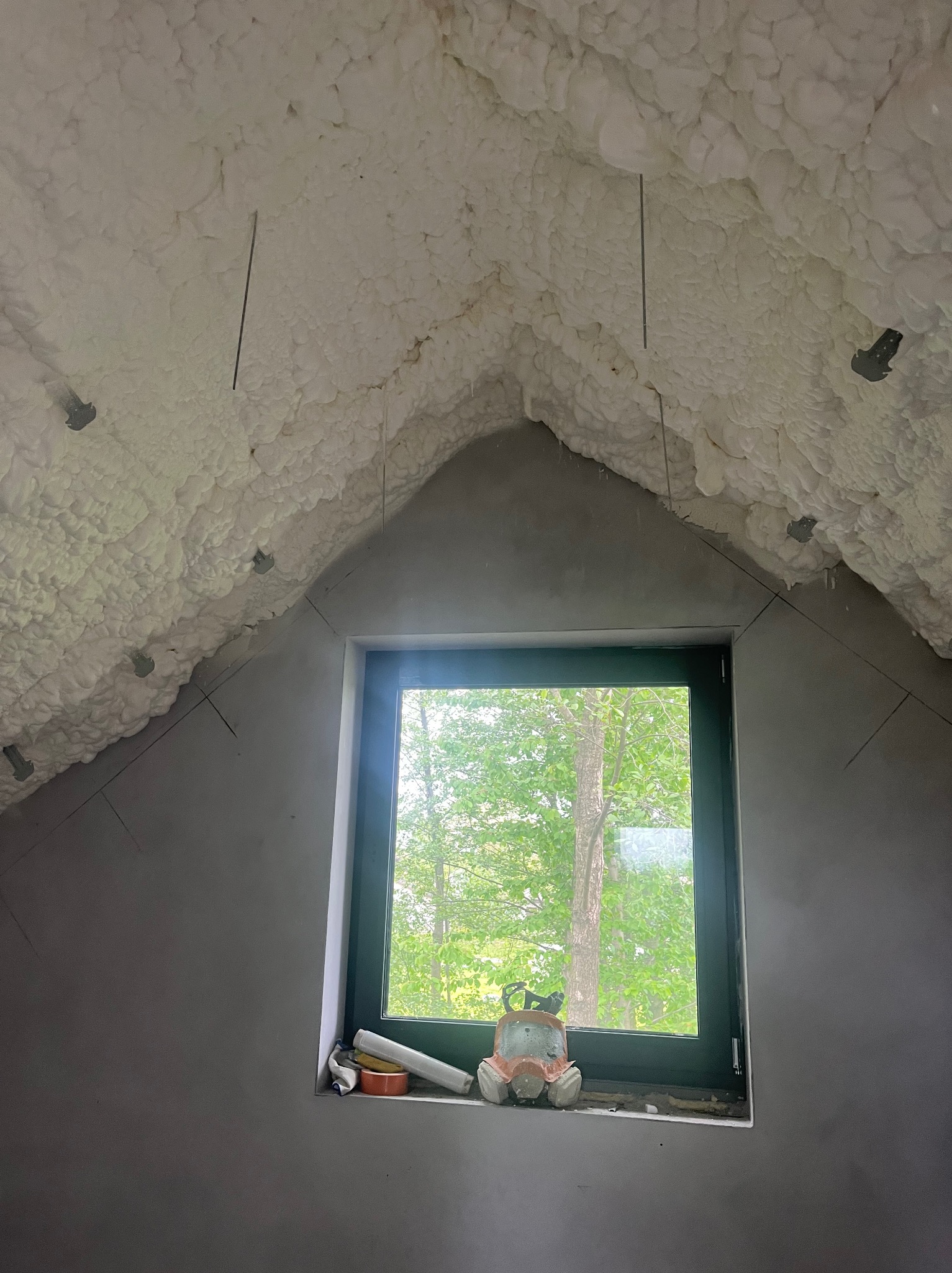 Poddasze po natrysku izolacji. Widok na okno z zielenią. Na parapecie leżą narzędzia pracy i maska ochronna. Izolacja pokrywa skosy dachu i sufit.