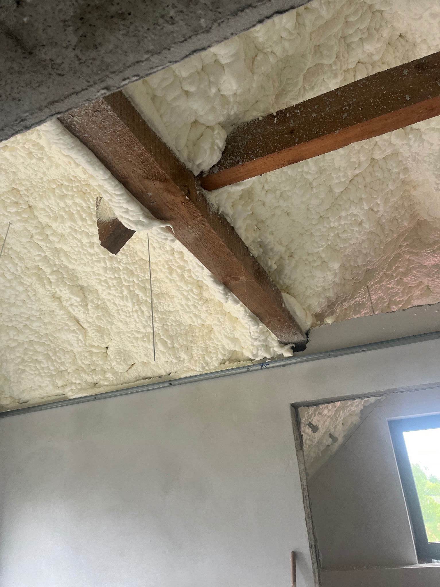 Ocieplenie poddasza pianką: drewniana belka w gęstej, białej izolacji, widok z dołu na fragment okna. Detal wykończenia.