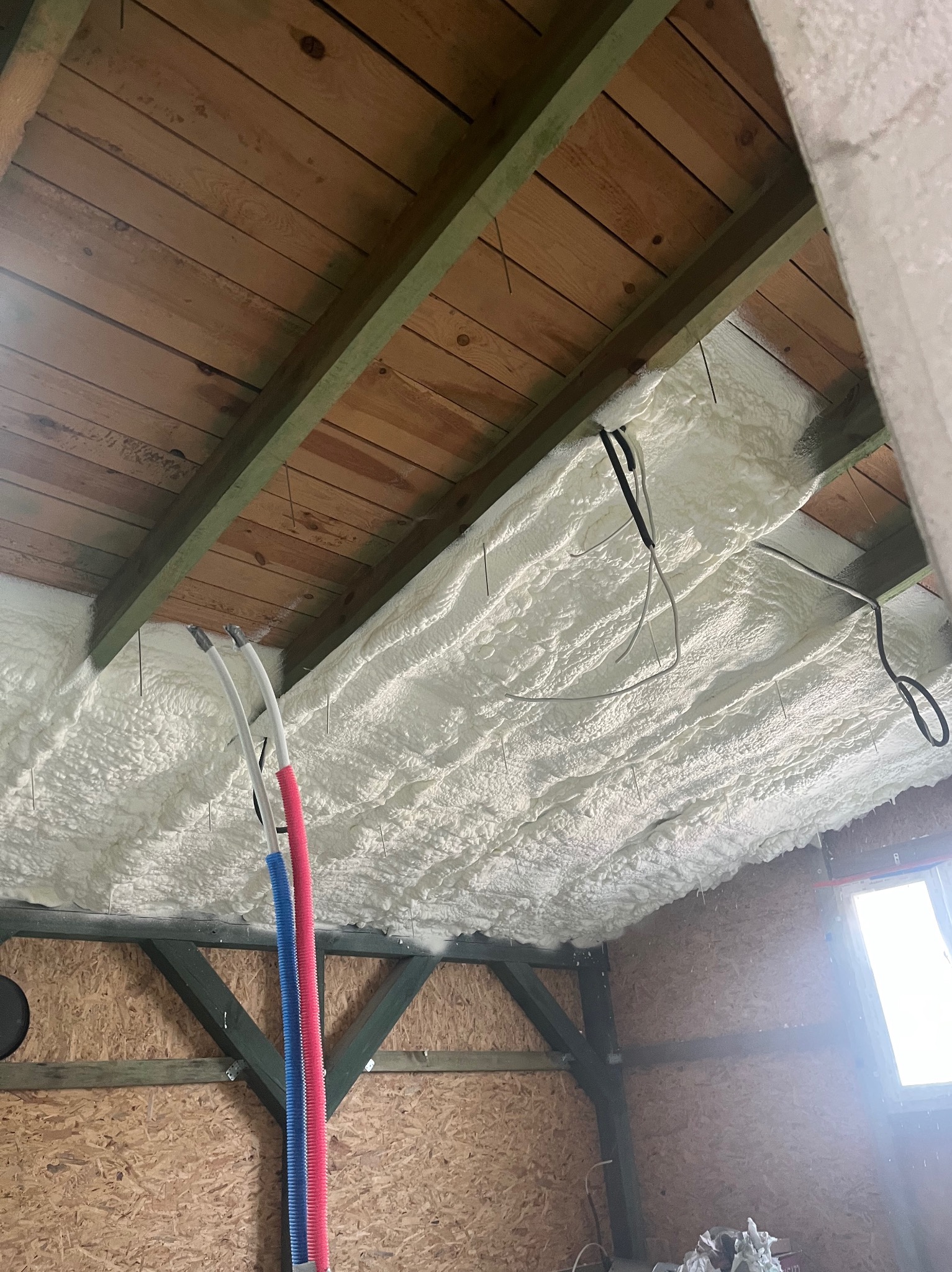 Ocieplenie poddasza pianką: biała, nierówna struktura wypełnia przestrzeń między drewnianymi belkami, widoczne przewody i fragment okna dachowego.