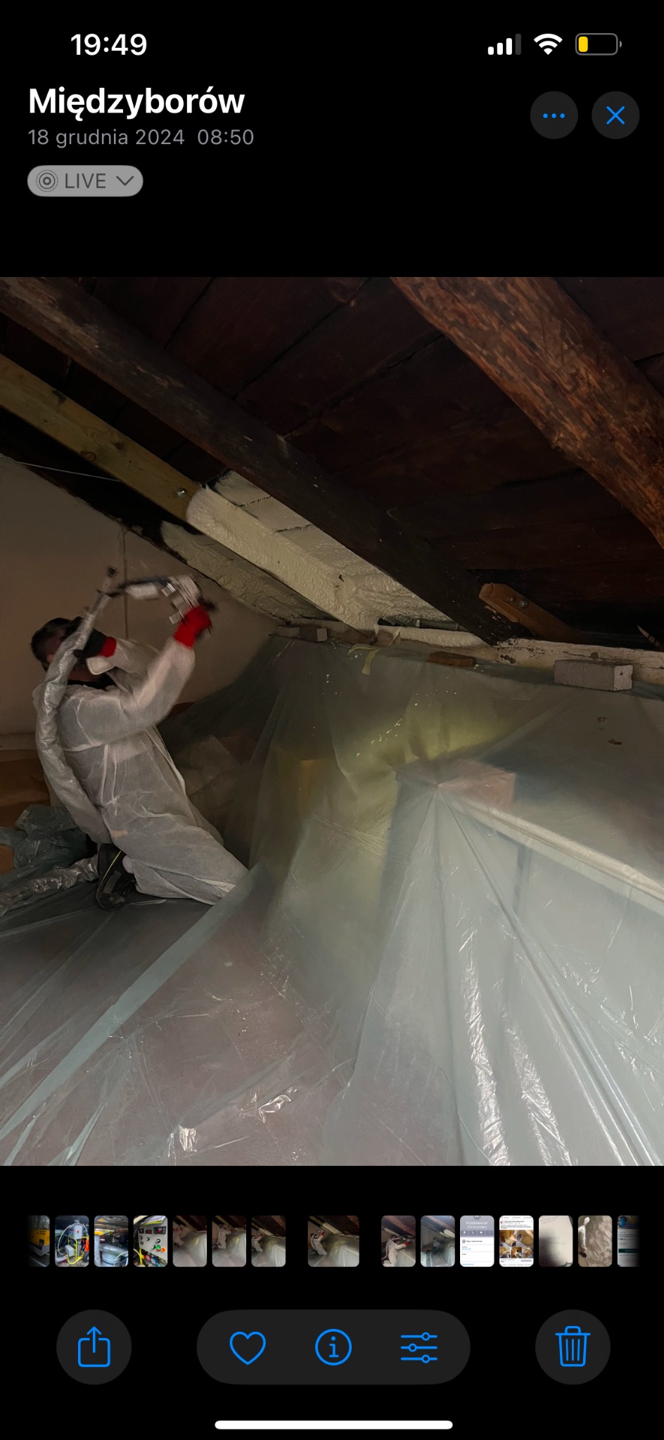 Pracownik w kombinezonie nanosi izolację piankową na poddasze, widoczne drewniane elementy konstrukcyjne i folia ochronna. Praca w trakcie realizacji.