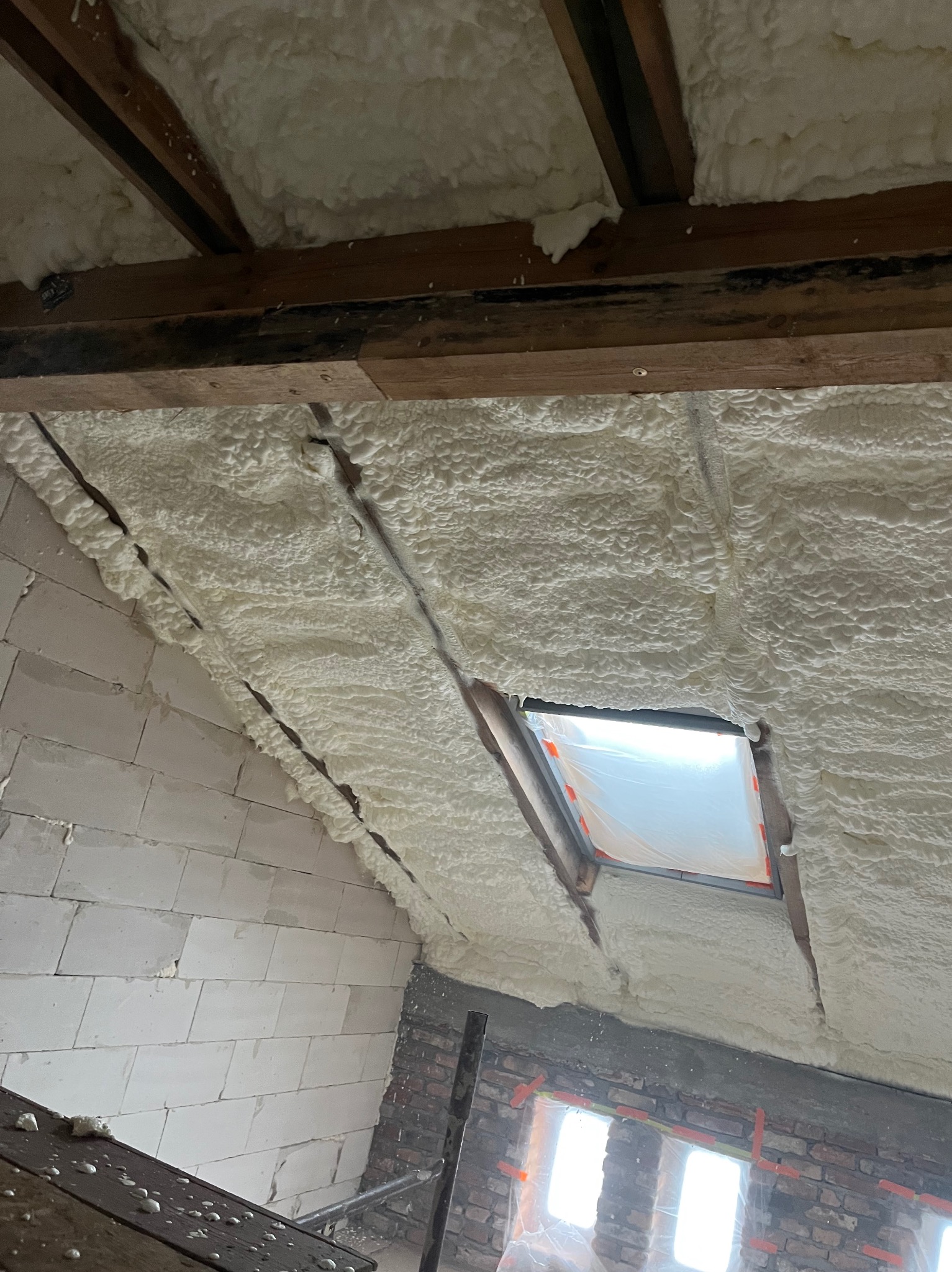 Poddasze po ociepleniu pianką PUR: widoczne drewniane belki, okno dachowe zabezpieczone folią i ściany z pustaków pokryte grubą warstwą pianki o strukturze gąbki.