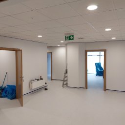 Michal Witowski - Wnętrze biura po malowaniu ścian na biało, z widocznymi otwartymi drzwiami, zabezpieczonymi listwami przypodłogowymi i sufit podwieszany z oświetleniem punktowym. W tle drabina i meble przykryte...