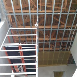 Michal Witowski - Montaż stelaża sufitu podwieszanego z płyty karton gips, widok z dołu, pracownik na rusztowaniu widoczny w tle.