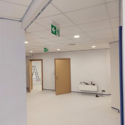 Michal Witowski - Wnętrze biura w Gnieźnie po świeżym malowaniu, z widocznymi taśmami malarskimi zabezpieczającymi krawędzie ścian, sufitu kasetonowego i podłogi, częściowo otwarte drewniane drzwi oraz drabina w tle.