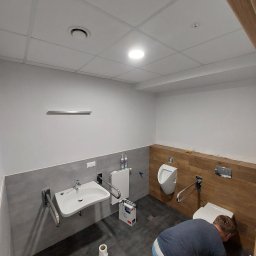 Michal Witowski - Pracownik na kolanach czyści podłogę w nowo wyremontowanej łazience z umywalką, toaletą i pisuarami przystosowanymi dla osób niepełnosprawnych, z widocznymi uchwytami pomocniczymi i wykończeniem...