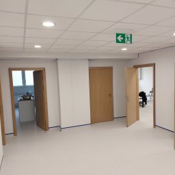 Michal Witowski - Wnętrze biurowe po remoncie, widoczne drzwi z ościeżnicami, podwieszany sufit z oświetleniem punktowym i lampą ewakuacyjną, zabezpieczone listwy przypodłogowe folią malarską.