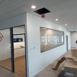 Michal Witowski - Wnętrze biura w trakcie wykańczania, widoczne ściany po szpachlowaniu, okna z widokiem na miasto, sufit kasetonowy z otworami na oświetlenie i instalacje, oraz materiały budowlane na podłodze.