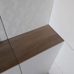 Fragment wykończonej łazienki z białymi płytkami o wzorzystej fakturze i drewnopodobnymi akcentami, widoczne fugi i narożnik, na podłodze folia ochronna.