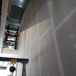 Wysoka ściana z płyt kartonowo-gipsowych, częściowo zaszpachlowana, widoczny dźwig budowlany z łańcuchem i hakiem, sufit z metalową konstrukcją w tle.