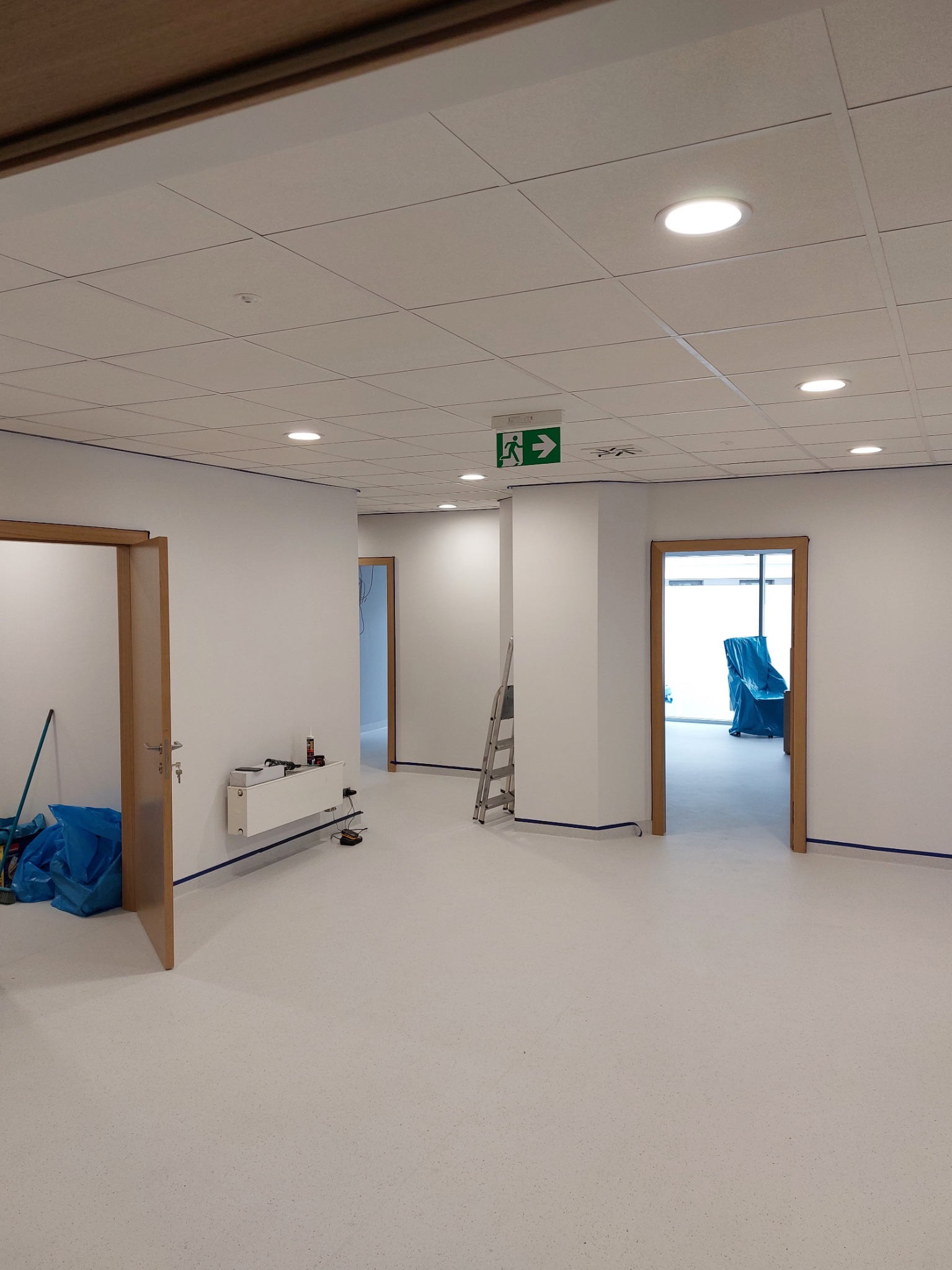 Wnętrze biura po malowaniu ścian na biało, z widocznymi otwartymi drzwiami, zabezpieczonymi listwami przypodłogowymi i sufit podwieszany z oświetleniem punktowym. W tle drabina i meble przykryte...