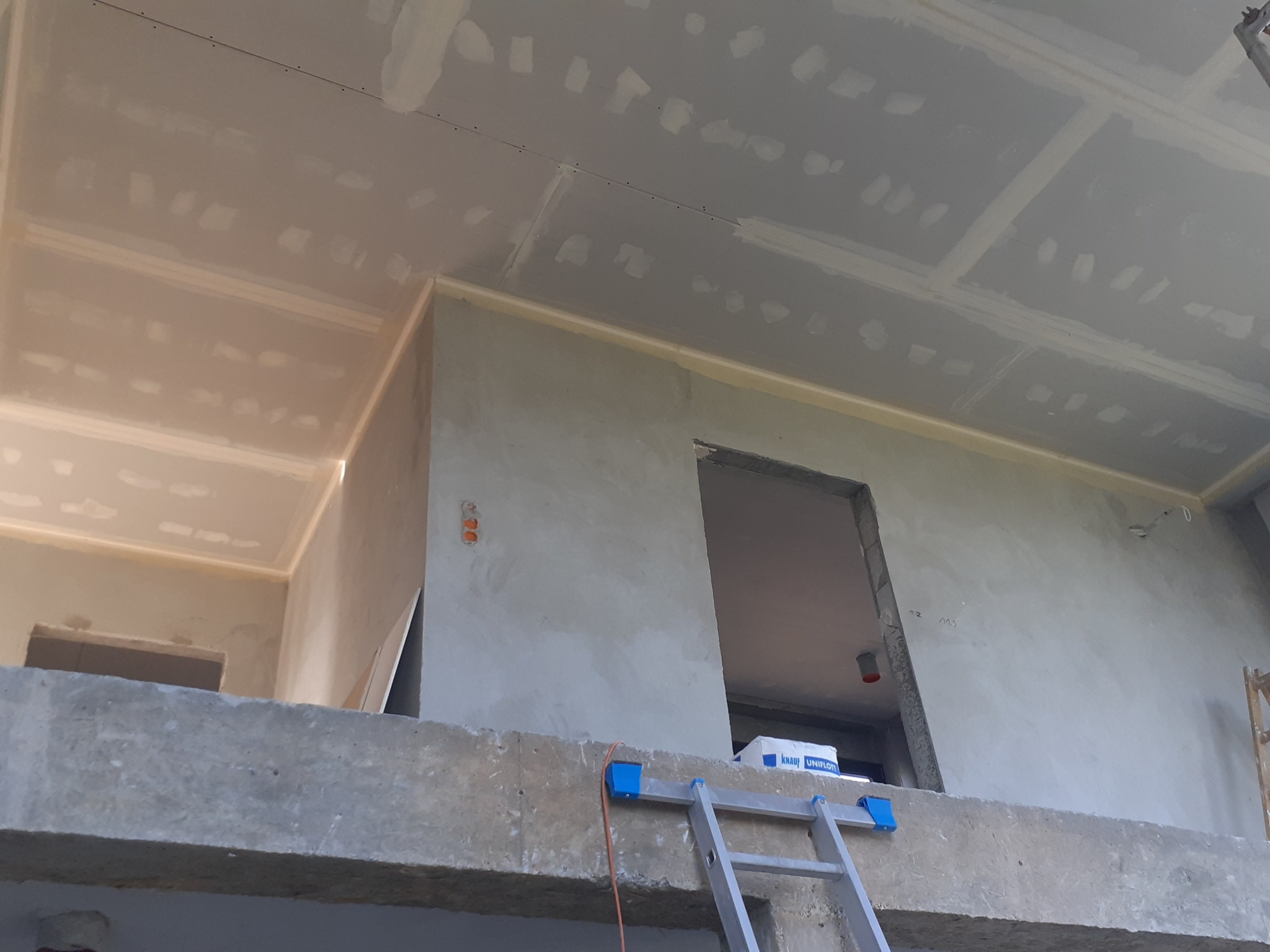 Wnętrze w trakcie wykańczania z widocznymi zamontowanymi płytami na suficie, częściowo zaszpachlowanymi, oraz ścianami przygotowanymi do dalszych prac, drabina oparta o betonową konstrukcję.
