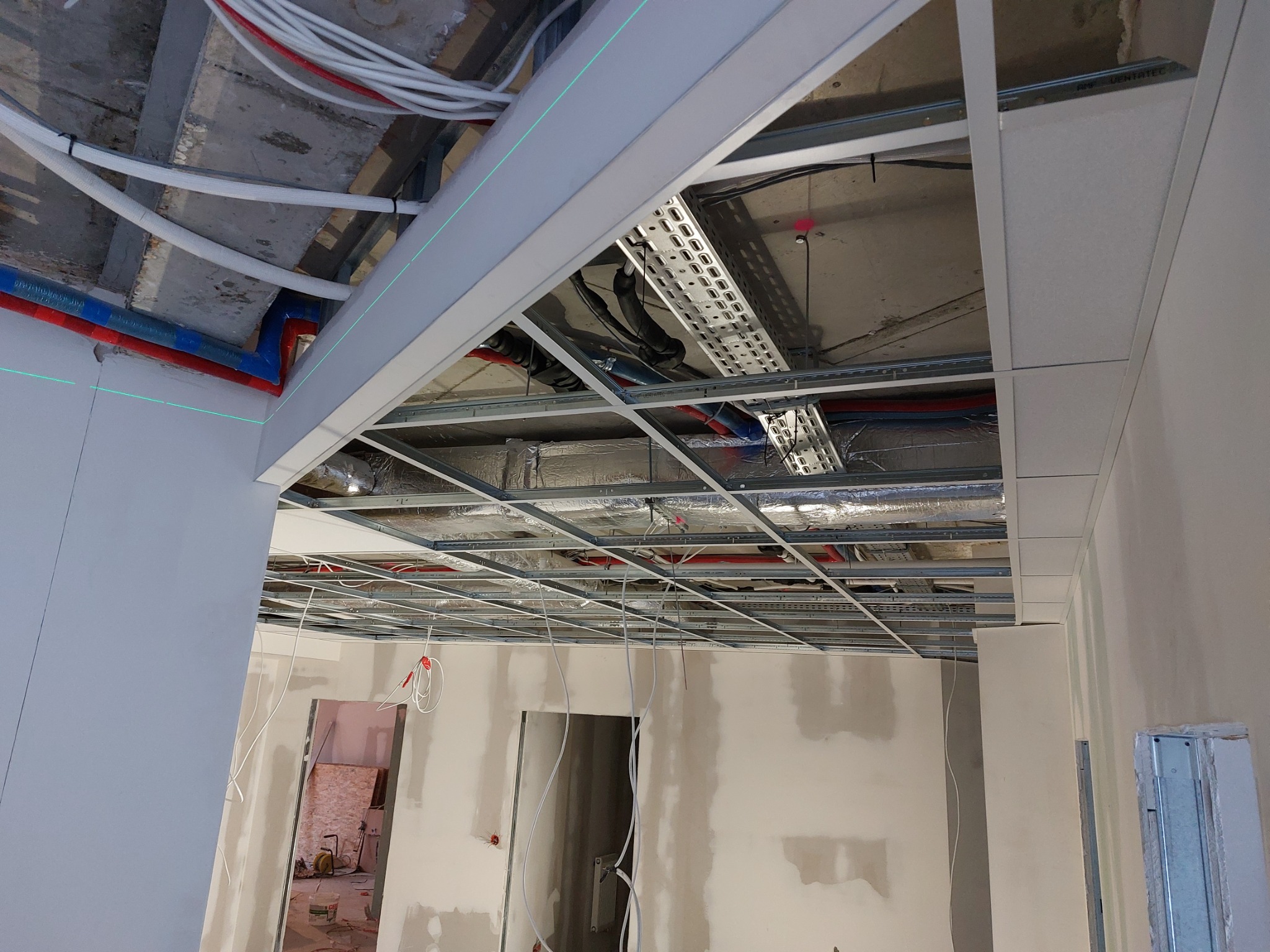 Montaż sufitu podwieszanego z widoczną konstrukcją metalową, instalacjami elektrycznymi i wentylacyjnymi, ściany w trakcie szpachlowania, zielona linia lasera wyznaczająca poziom.