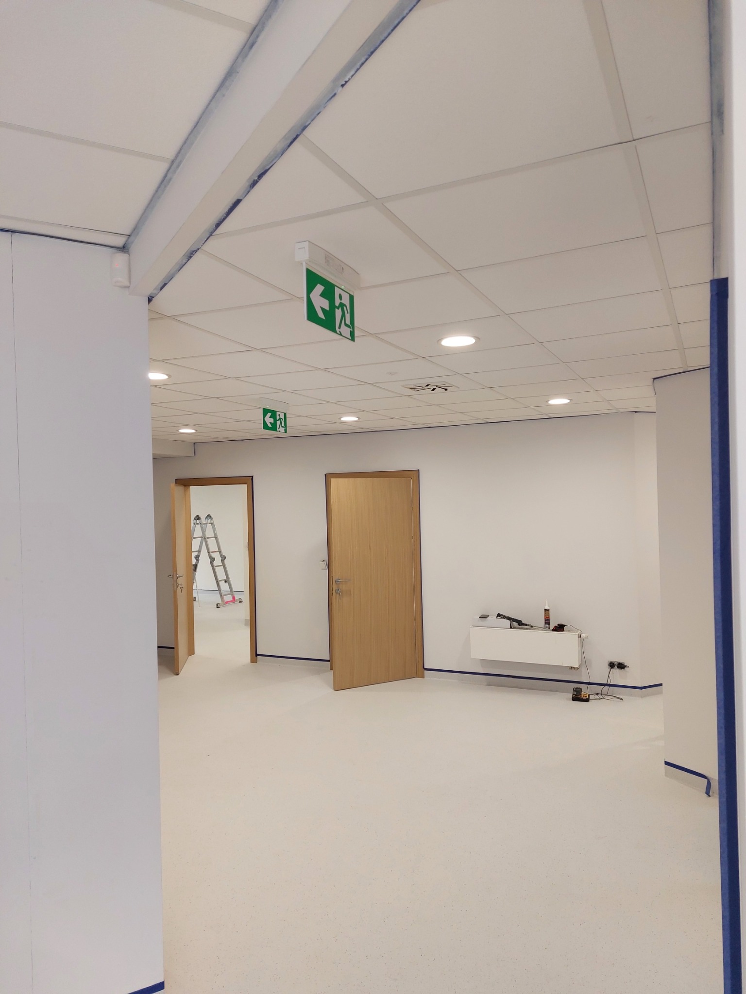 Wnętrze biura w Gnieźnie po świeżym malowaniu, z widocznymi taśmami malarskimi zabezpieczającymi krawędzie ścian, sufitu kasetonowego i podłogi, częściowo otwarte drewniane drzwi oraz drabina w tle.