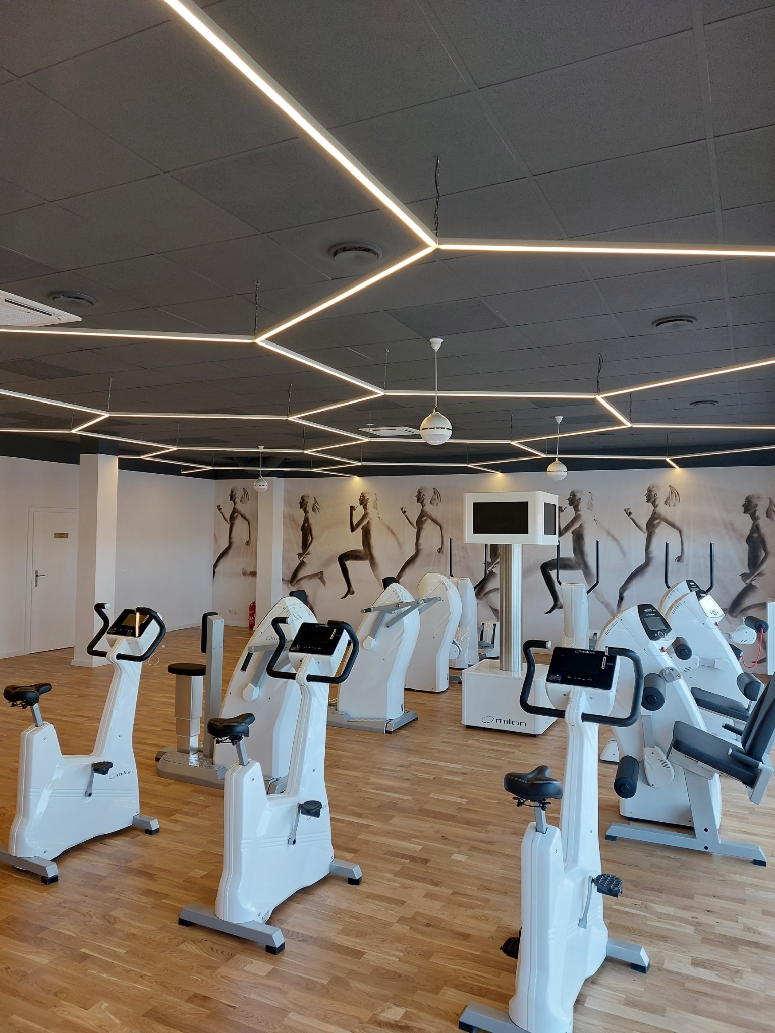 Wnętrze jasnej sali fitness z białymi maszynami do ćwiczeń, drewnianą podłogą i sufitem z geometrycznym oświetleniem LED. Na ścianie widoczna grafika przedstawiająca sylwetki biegnących kobiet.