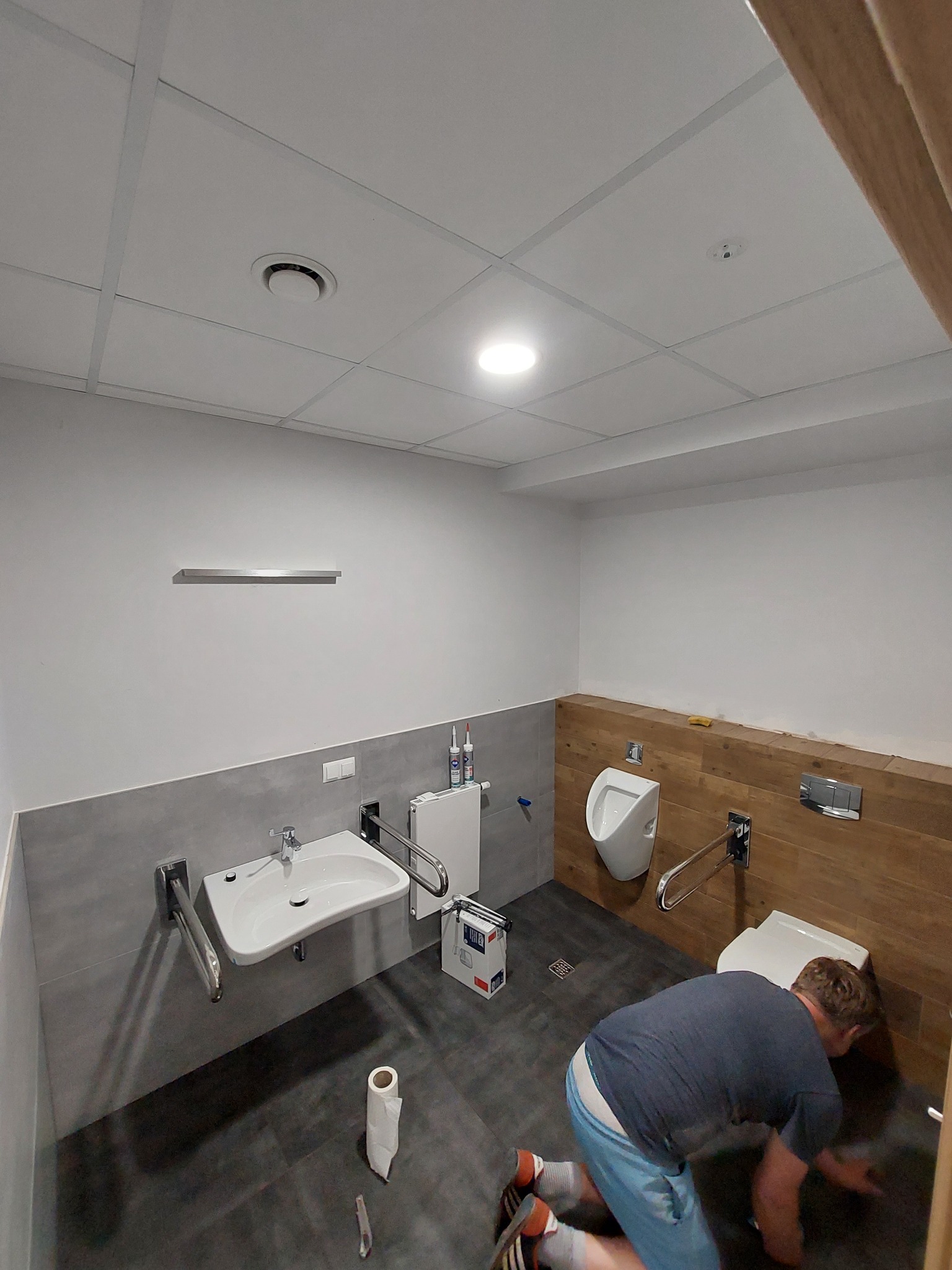 Pracownik na kolanach czyści podłogę w nowo wyremontowanej łazience z umywalką, toaletą i pisuarami przystosowanymi dla osób niepełnosprawnych, z widocznymi uchwytami pomocniczymi i wykończeniem...