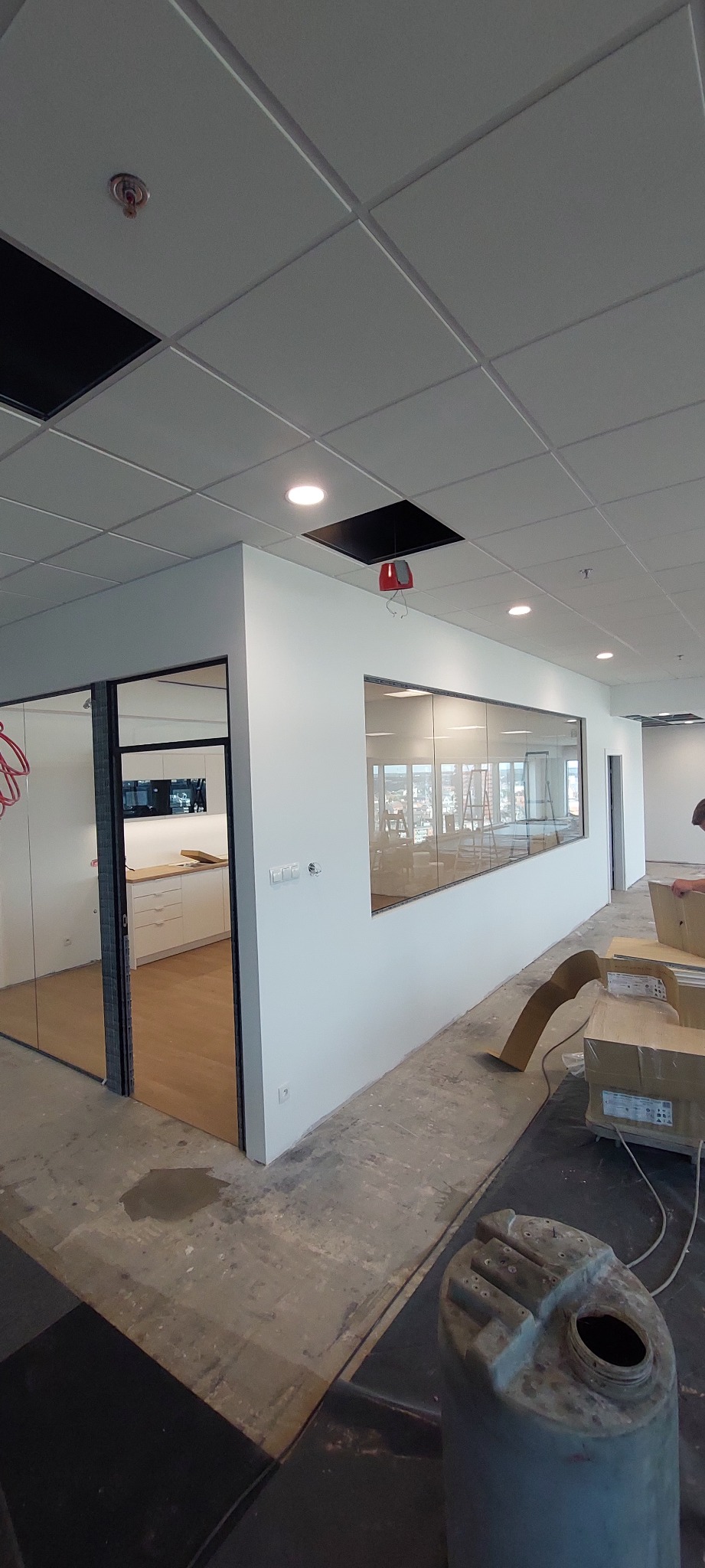Wnętrze biura w trakcie wykańczania, widoczne ściany po szpachlowaniu, okna z widokiem na miasto, sufit kasetonowy z otworami na oświetlenie i instalacje, oraz materiały budowlane na podłodze.