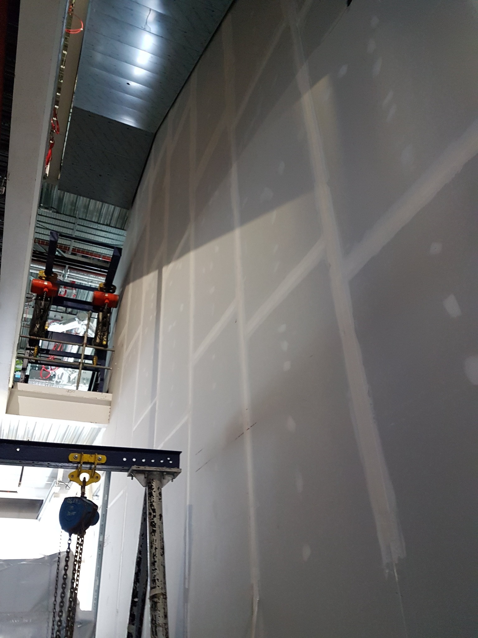 Wysoka ściana z płyt kartonowo-gipsowych, częściowo zaszpachlowana, widoczny dźwig budowlany z łańcuchem i hakiem, sufit z metalową konstrukcją w tle.