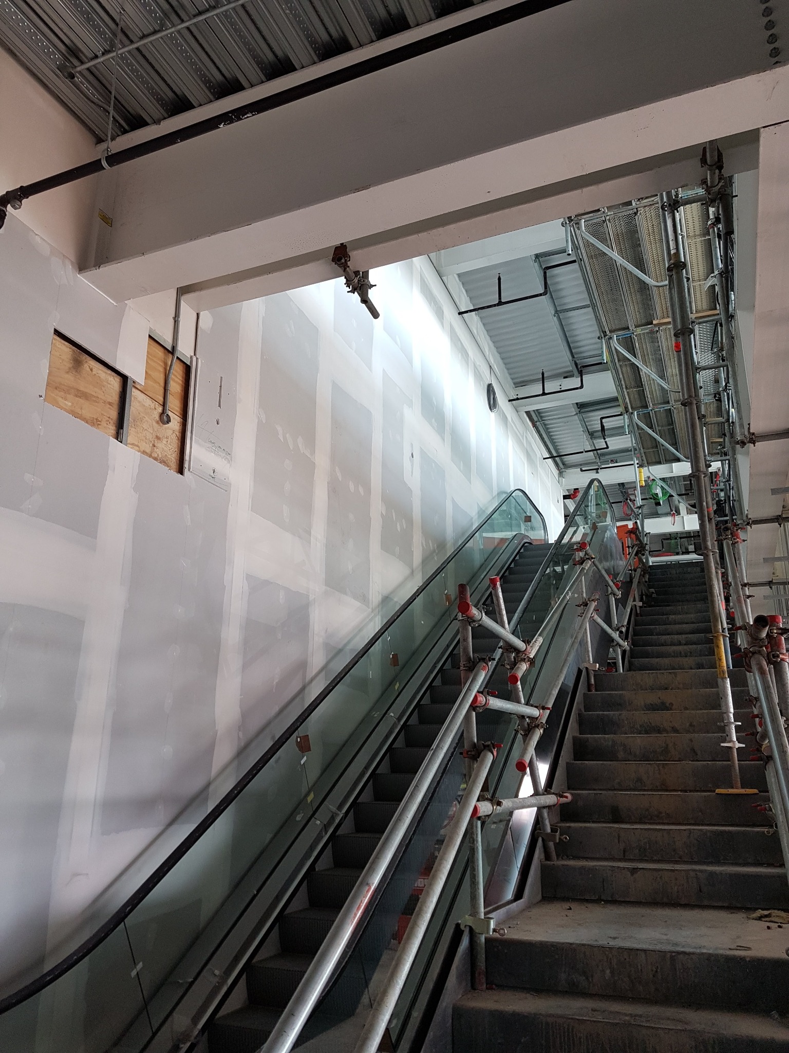 Wnętrze w trakcie remontu z widocznymi schodami ruchomymi, ścianami pokrytymi płytami gipsowo-kartonowymi przed wykończeniem, oraz rusztowaniami zabezpieczającymi prace na wysokościach.