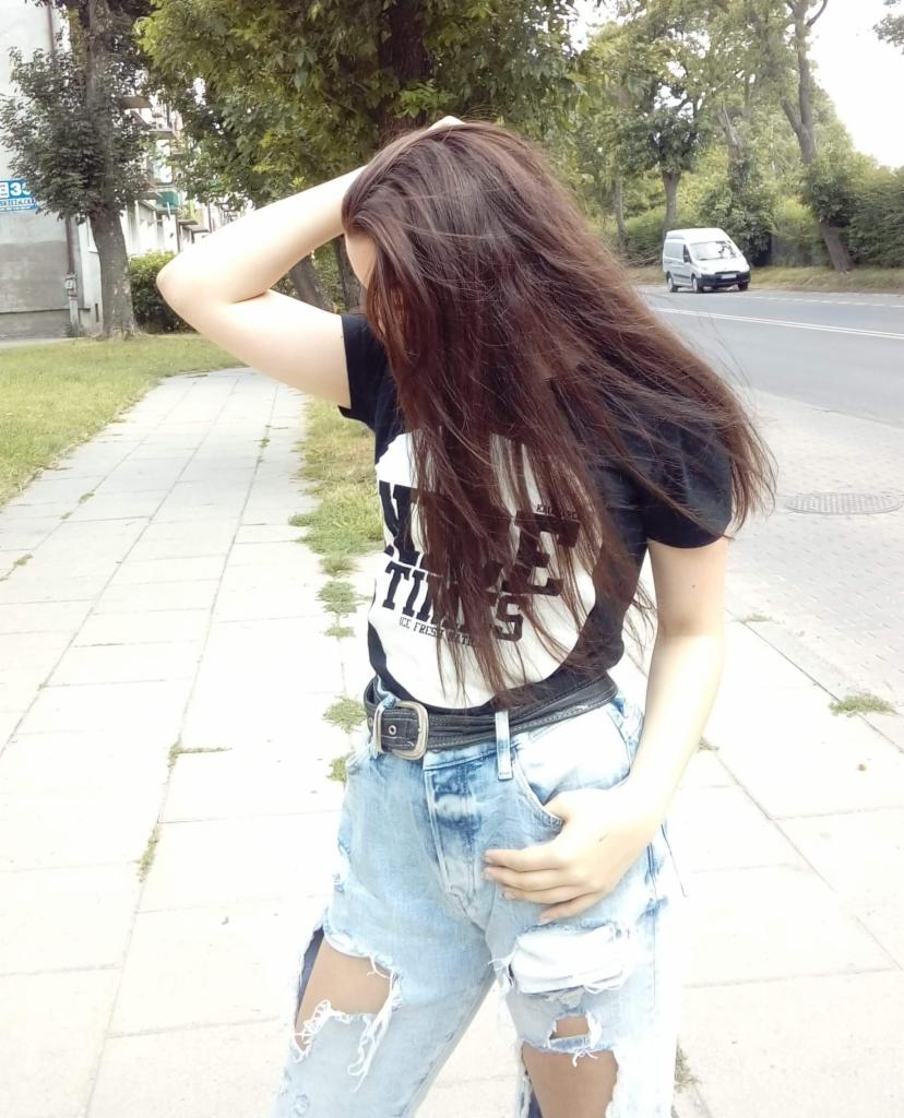 Kobieta w podartych jeansach i czarnej koszulce stoi na chodniku, z dłonią na włosach. W tle ulica i budynek.