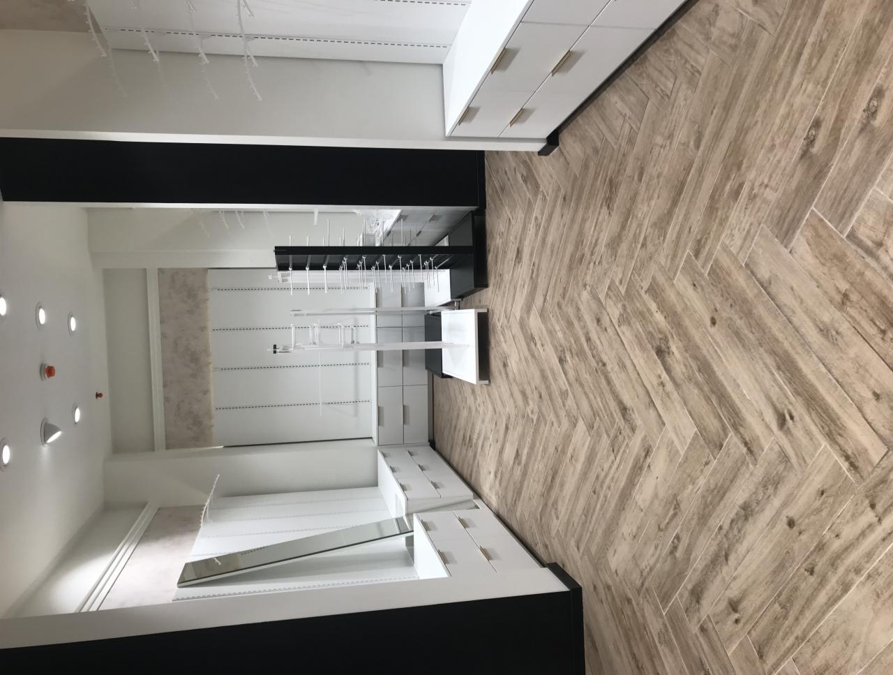 Jasne, nowocześnie urządzone wnętrze sklepu z drewnopodobną podłogą ułożoną w jodełkę, białymi meblami i oświetleniem punktowym.