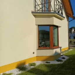 Nowoczesna balustrada balkonowa o geometrycznym wzorze, wykonana z ciemnego metalu, zamontowana przy oknie wykuszowym w jasnym budynku z żółtą podmurówką i ozdobnym obrzeżem z białego kamienia...