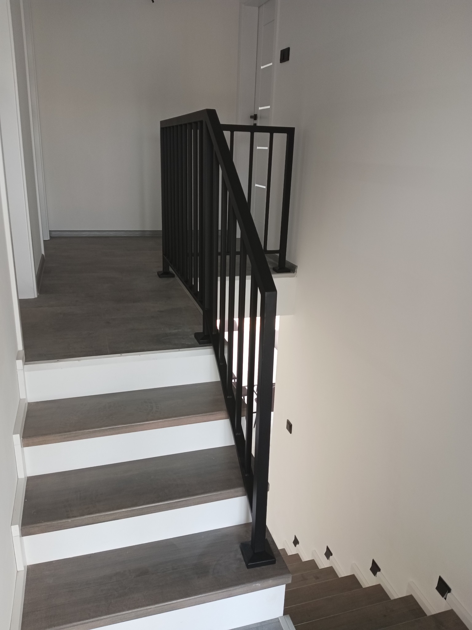 Nowoczesna, czarna metalowa balustrada schodowa z pionowymi szczeblami, zamontowana na schodach z drewnopodobnymi stopniami i białymi podstopnicami, widok z góry.