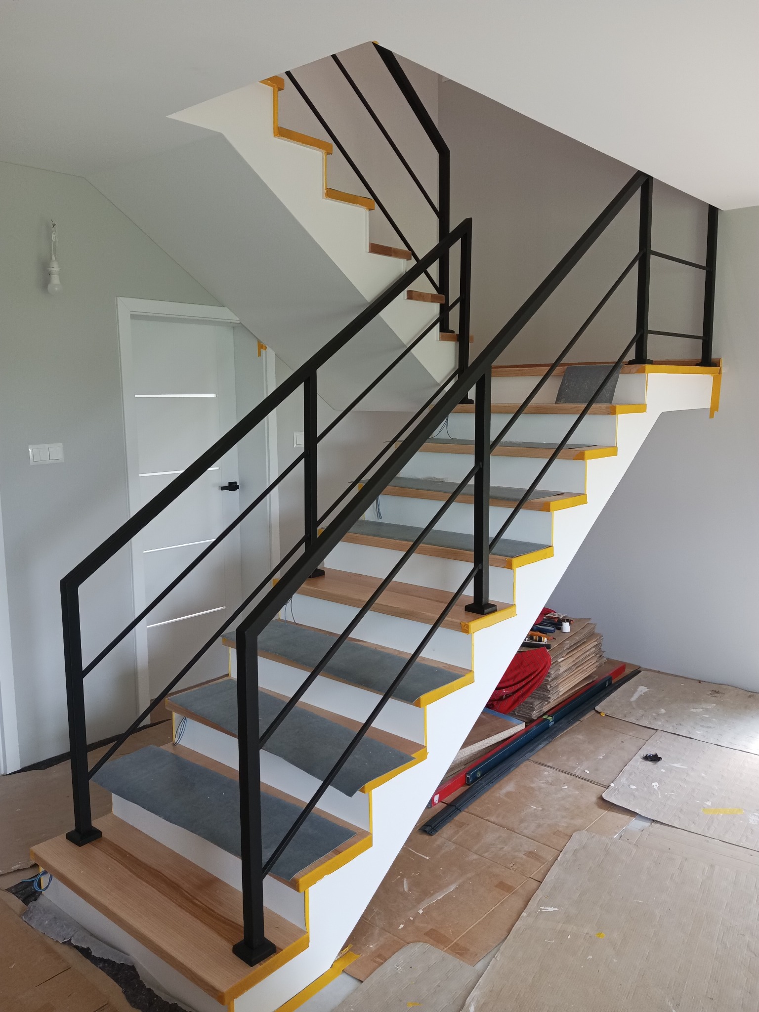 Nowoczesne schody z czarną, metalową balustradą, drewnianymi stopniami i białymi podstopnicami z żółtymi akcentami. Widoczne zabezpieczenia podczas montażu.