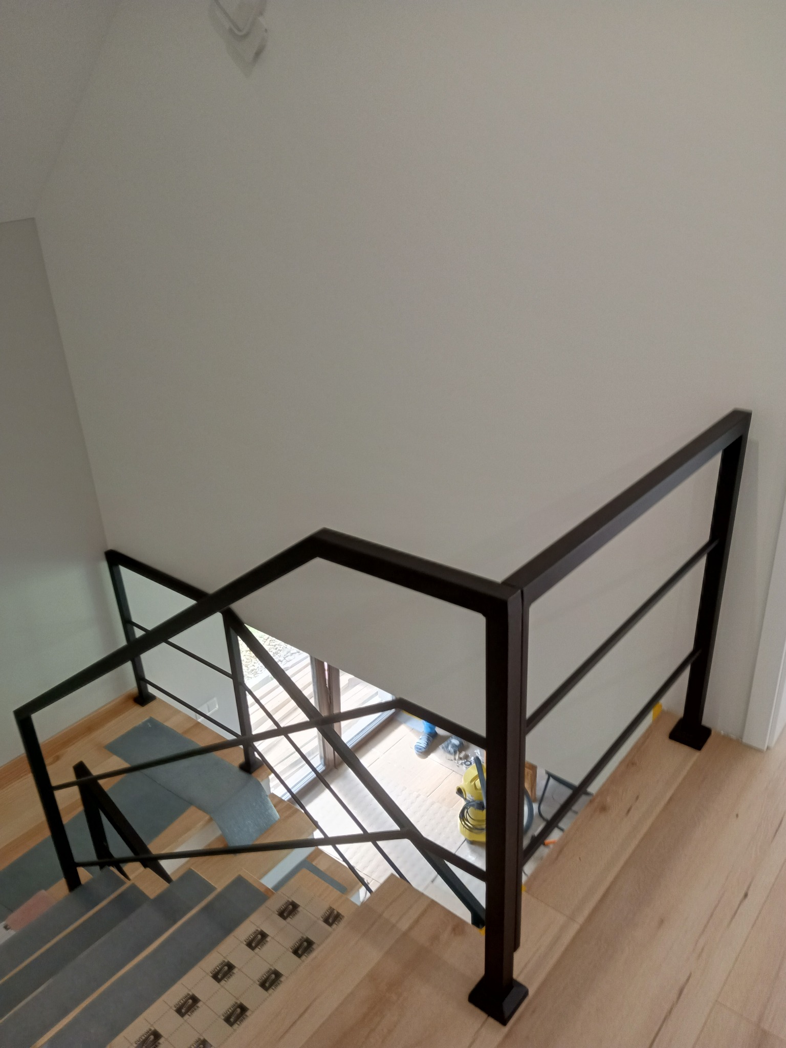 Nowoczesna, czarna balustrada na schodach z drewnianymi stopniami. Widok z góry, częściowo widoczne narzędzia pracy, wnętrze domu w trakcie wykończenia.