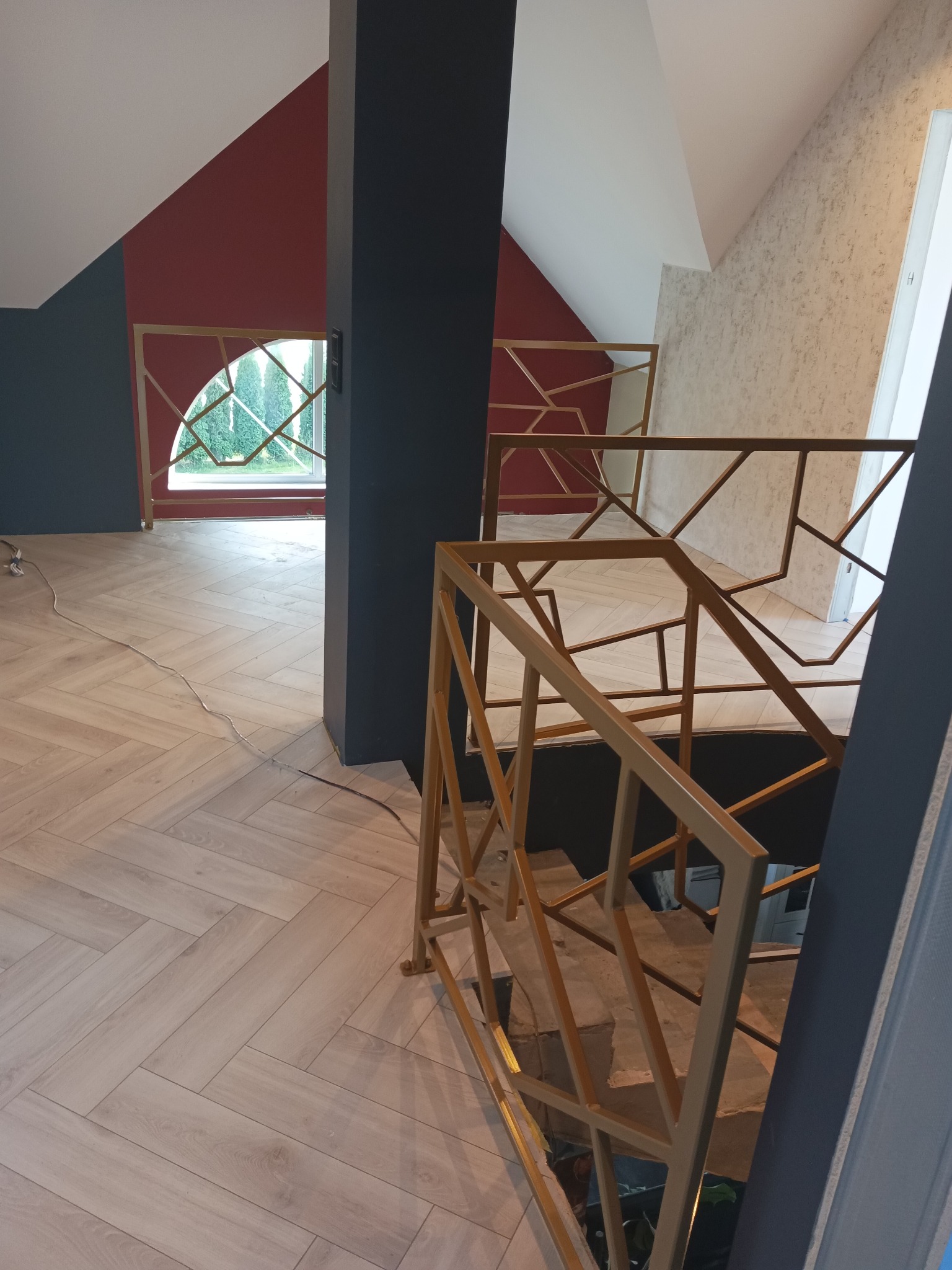 Nowoczesna, złota balustrada schodowa z geometrycznym wzorem, w jasnym wnętrzu z parkietem w jodełkę i oknem z widokiem na zieleń. Nietypowy design.