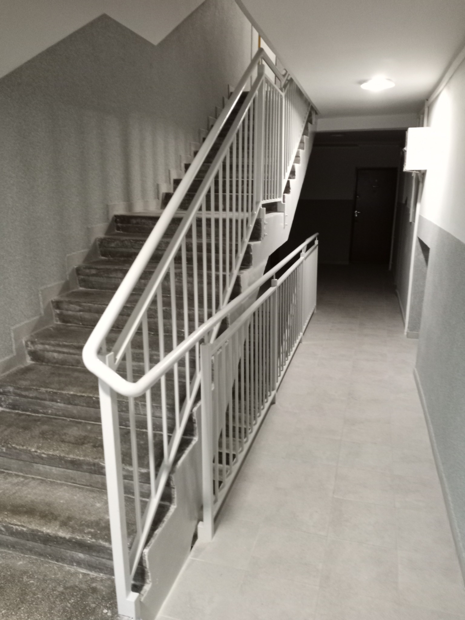 Klatka schodowa w bloku z białymi metalowymi poręczami i schodami o szarych stopniach, widok z dołu na górę.