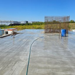 CONTEAM sp. z o.o. - Wylany betonowy fundament hali z wystającymi prętami zbrojeniowymi i konstrukcją zbrojeniową w tle, przyczepy budowlane w oddali, słoneczny dzień.