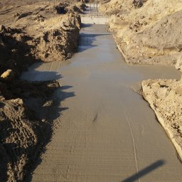 CONTEAM sp. z o.o. - Wylany fundament pod budynek z wystającymi prętami zbrojeniowymi, otoczony wykopem i ziemią. W tle widoczny budynek z żółto-czerwonymi panelami.