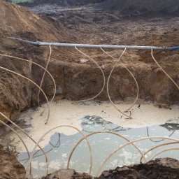 CONTEAM sp. z o.o. - Odwadnianie wykopu pod budowę: system rur zanurzonych w wodzie, podłączonych do głównej rury odprowadzającej wodę. Grunt gliniasty, widoczne ślady po wykopie.