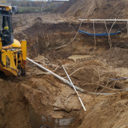 CONTEAM sp. z o.o. - Żółta koparka przy wykopie z rurami i kablami. Widoczne warstwy ziemi i elementy konstrukcyjne. Praca w toku, przygotowanie terenu pod budowę.
