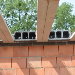 Wznoszenie ściany z czerwonej cegły ceramicznej, z widocznymi szarymi bloczkami betonowymi i elementami stropu Teriva, zieleń drzew w tle.